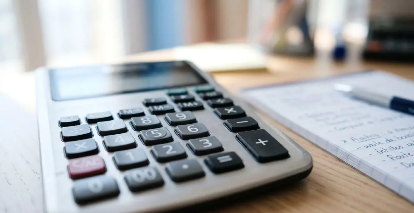 Gros plan sur une calculatrice posée sur un bureau blanc épuré avec un cahier de notes manuscrites floues en arrière-plan